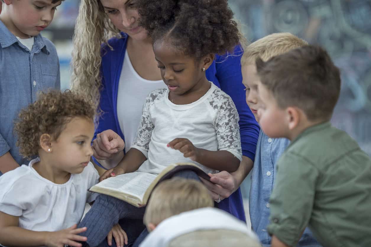 Five children of different ethnicity looking at a bible being held by an adult woman who is wearing a white top and blue cardigan.