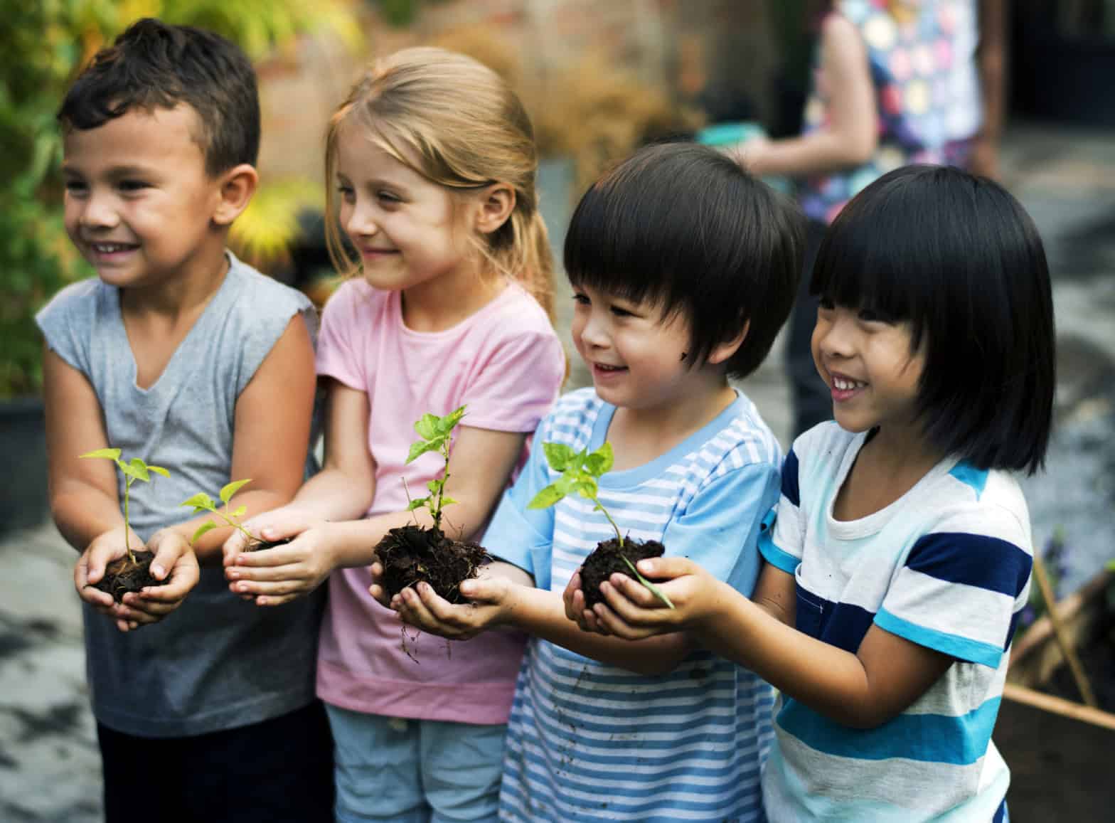 A group of diverse children in t-shirts outside holding small sapling plants