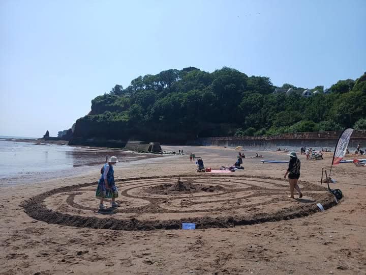 A labyrinth circle in sand on a beath with people making it. A dog is near the centre.