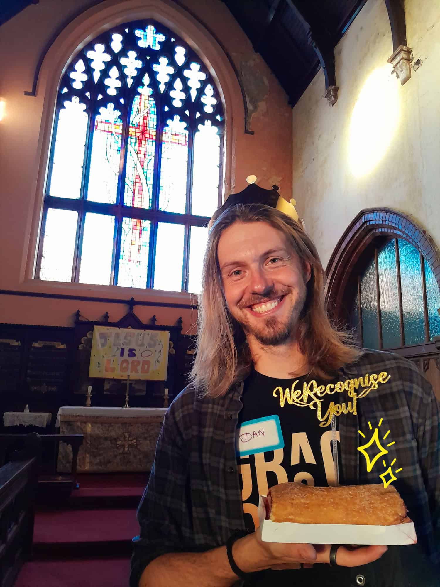 A young man wearing a name badge which says Dan is holding a cake and smiling whilst in Church.