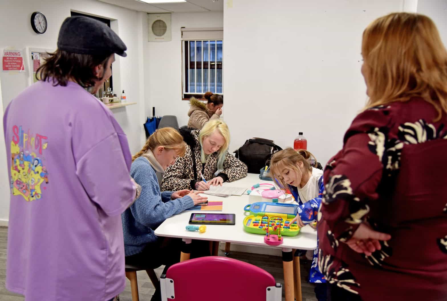 A community setting with adults and children sitting at a table with colouring activities