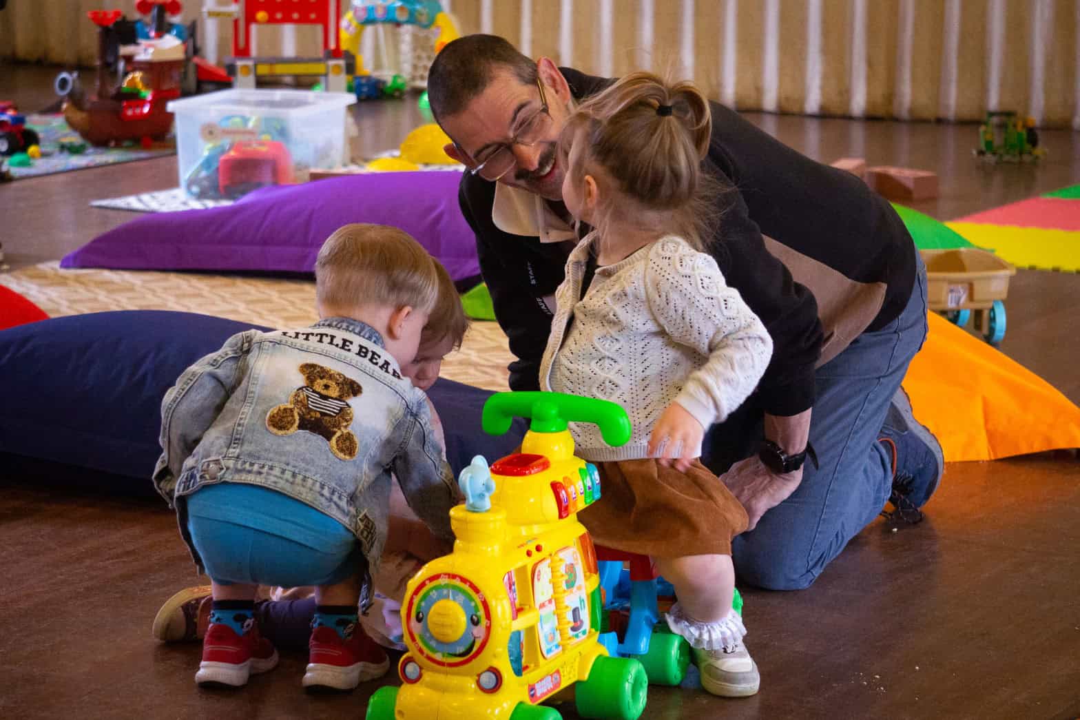 Two young children on play equipment in a community setting with an adult male interacting with them both.