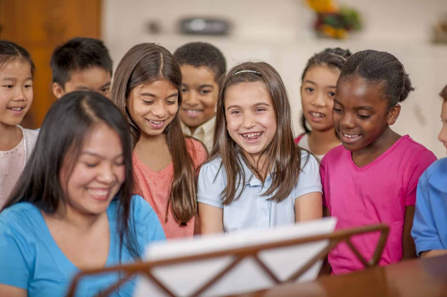 A group of diverse children standing around a piano. A female teacher is playing it whilst the children smile and get ready to sing in a choir.
