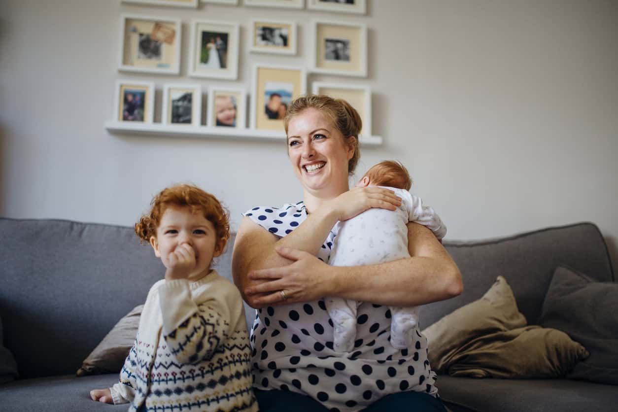 Mother with her two children in the living room of their home. The mother is holding a baby and the other little girl is laughing and looking at the camera while sucking her thumb.