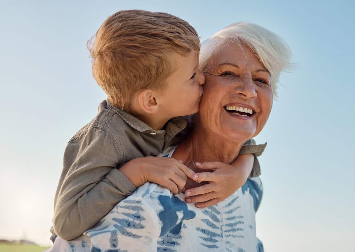 A Grandmother carrying her young grandson on her back whilst smiling. Her grandson is kissing her.