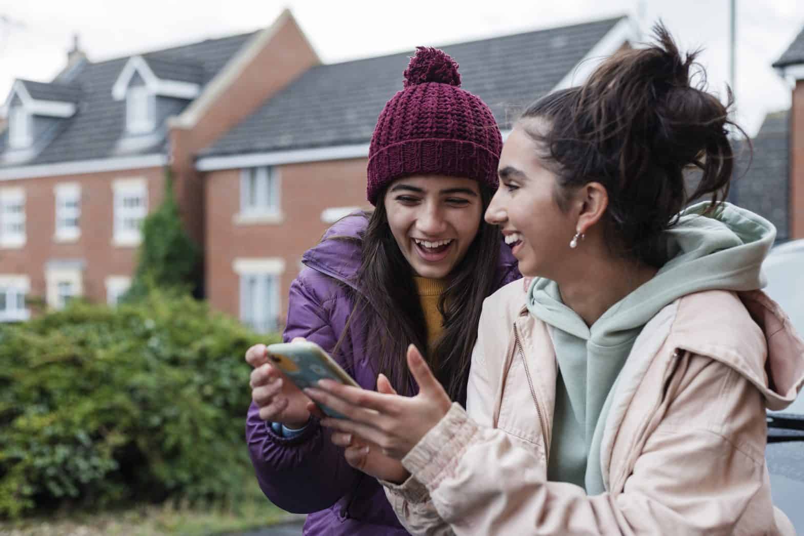 Two young teenage girls are outside a house laughing and smiling whilst looking at a Smartphone